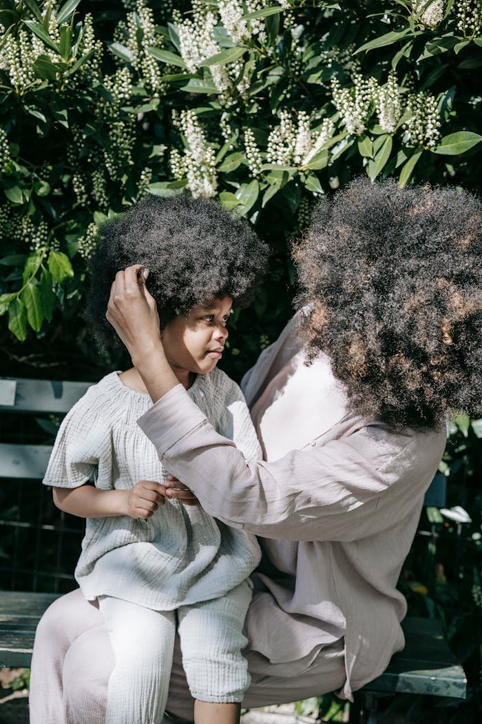 A Person in White Long Sleeve Shirt Touching the Hair of a Girl Sitting on her Lap